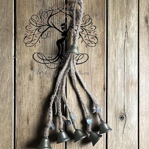 Könnte beinhalten: Ein rustikaler Windspiel mit mehreren kleinen Glocken im antiken Stil, die an geflochtenem Bindfaden aufgehängt sind. Die Glocken sind in verschiedenen Größen gehalten und hängen vor einem verwitterten Holzgrund. Der Hintergrund zeigt auch ein dekoratives Baumdesign.