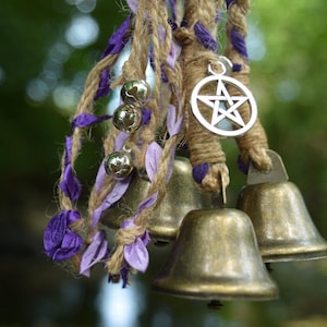 May include: A hanging decoration featuring three brass-coloured metal bells suspended by braided jute twine interwoven with purple and light purple fabric ribbons. A silver-toned metal pentagram charm and several small silver jingle bells are attached.