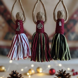 Può includere: Tre ornamenti a nappa fatti a mano appesi a un ramo. Ogni ornamento ha una testa intrecciata, un corpo bordeaux e una gonna a nappa in diversi colori: rosso e bianco, rosso e marrone e nero e verde. Sono attaccati ciondoli d'argento. Decorazioni natalizie.