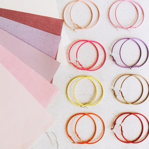 May include: Assortment of ten colorful hoop earrings in various sizes and colors, including pink, purple, yellow, and orange. The earrings are arranged on a white surface.