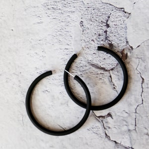 May include: A pair of matte black hoop earrings featuring a circular design with a smooth finish and thin silver-toned metal posts for closure. The earrings are simple, circular, and have a consistent thickness throughout the entire hoop structure.
