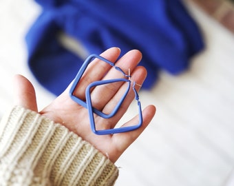 May include: A pair of matte blue hoop earrings with a distinct square shape. The earrings feature a smooth, solid blue finish and a standard post-back closure mechanism, designed as a geometric accessory with clean lines and a bold, vibrant color.