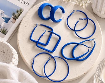 May include: A collection of five pairs of bright blue earrings in various shapes, including thick small hoops, thin large circular hoops, square-shaped hoops, and elongated oval hoops, all arranged on a circular textured stone surface.