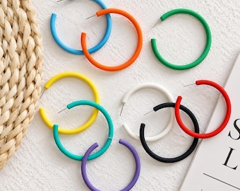 May include: A collection of nine circular hoop earrings in various solid colors including blue, orange, green, yellow, teal, purple, white, black, and red. Each earring features a thin metal post for attachment, arranged in a scattered pattern.