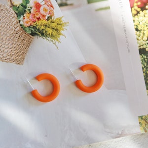 May include: A pair of orange hoop earrings. The earrings are made of a thick, rounded material and have a simple, modern design.