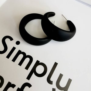 May include: A pair of matte black hoop earrings with a thick, rounded profile and silver-toned metal posts. The earrings are positioned on a white surface featuring the partial black text Simply perf, which is clearly visible in the background.