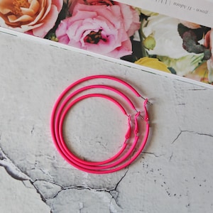 May include: A pair of bright neon pink hoop earrings featuring a thin, circular metal design. The earrings are constructed with a simple post-back closure mechanism, showcasing a vibrant, solid color finish that stands out clearly in this view.
