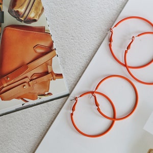 May include: Three pairs of large, orange hoop earrings. The earrings are made of a thick, smooth material and have a simple, modern design.