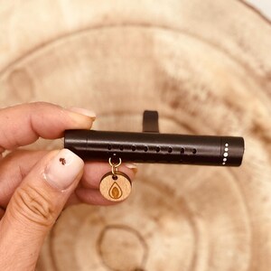 Puede incluir: Ambientador de coche negro con un amuleto de madera con un diseño de hoja. El amuleto está unido al ambientador con una cadena dorada.