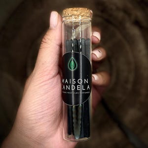 Könnte beinhalten: Ein klares Glasfläschchen mit Korken, gefüllt mit schwarzen Räucherstäbchen. Das Fläschchen hat ein schwarzes Etikett mit dem Text "MAISON ANDELA" und einem grünen Blattlogo. Der Text "IONS VEGETALES PARFUMEES" ist ebenfalls vorhanden.