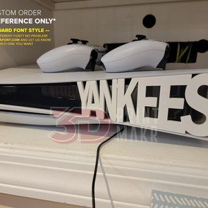 May include: White gaming console with two white controllers. The console has the word "Yankees" cut out of the front. The text "CUSTOM ORDER" and "STANDARD FONT STYLE" is also visible.