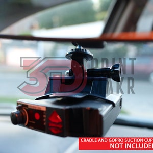 May include: A black 3D printed mount for a radar detector, attached to a rearview mirror in a car. The mount has a GoPro-style attachment point. "CRADLE AND GOPRO SUCTION CUP MOUNT/SCREW NOT INCLUDED" is printed on the bottom of the image.