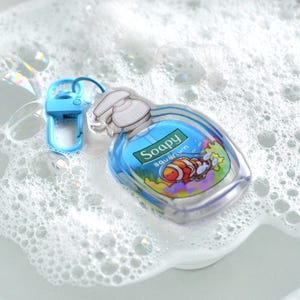 May include: A clear, soap-shaped keychain with a blue liquid interior and a white pump top. The label reads "Soapy aquarium" and features a cartoon clownfish. A small, blue clip is attached to the keychain.