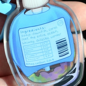 May include: A clear, blue-tinted acrylic charm in the shape of a bottle. The charm features a label with the text "Ingredients: soap, clownfish, coral reef, the entire ocean, whimsy, childlike wonder." The bottom of the label reads "*may contain fish*".
