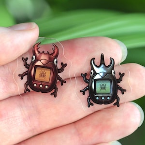 May include: Two transparent stickers depicting beetle-shaped digital pet toys. One is brown with red details, the other is black. Each has a small screen showing a pixelated graphic and three buttons.