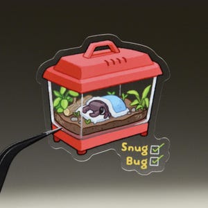 May include: A die-cut sticker featuring a red plastic pet carrier containing a brown beetle sleeping under a light blue blanket on brown soil with green plants and a small log. The text Snug Bug is printed in yellow with two green checkmarks.