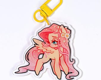 Puede incluir: Un llavero acrílico transparente con un pony de dibujos animados con pelo rosa, una corona de flores y alas. El pony tiene un cuerpo de color canela claro y está unido a un cierre de metal amarillo.