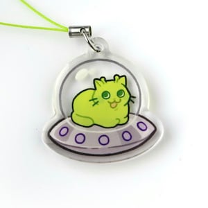 May include: A clear acrylic charm depicting a lime green cat in a spaceship. The cat is smiling and sits on a purple and grey saucer. The charm has a silver clasp and a green cord.