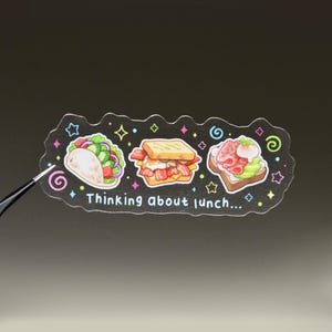 May include: A black sticker with a scalloped edge featuring illustrations of a taco, a bacon sandwich, and avocado toast. The text "Thinking about lunch..." is printed in white, surrounded by colourful stars and swirls on a dark background.