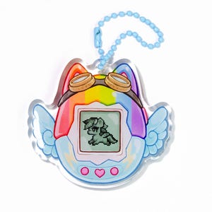 Op de afbeelding: Een heldere acryl bedel in de vorm van een retro handheld game, met een regenboogontwerp en een gepixeld pony-personage op het scherm. Het heeft vleugels, een bril en een blauwe kogelketting voor bevestiging.