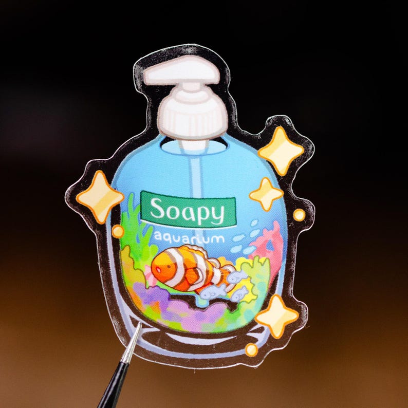 May include: A sticker in the shape of a soap dispenser with a blue background and the words "Soapy aquarium." The sticker features a cartoon clownfish, coral, and bubbles. Yellow star accents surround the dispenser.