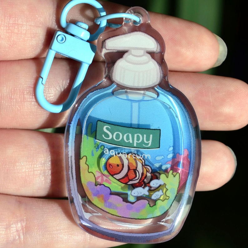 May include: A clear acrylic keychain shaped like a soap bottle, featuring a cartoon clownfish and the text "Soapy aquarium." The keychain has a blue clasp and a blue top, with a colorful underwater scene inside.