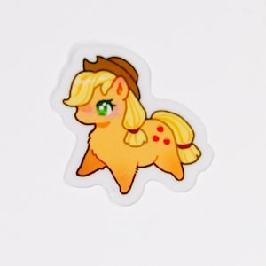 May include: A cartoon sticker of a pony with a light brown hat, yellow hair, and a yellow body. The pony has green eyes and three red apple markings on its rear. The sticker has a white border.