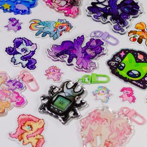 May include: A selection of vibrant acrylic keyrings and stickers showcasing cartoon characters and designs. The keyrings have metal clasps in various colours. The stickers include a green cat with the word "mrow" and a beetle with a digital screen.