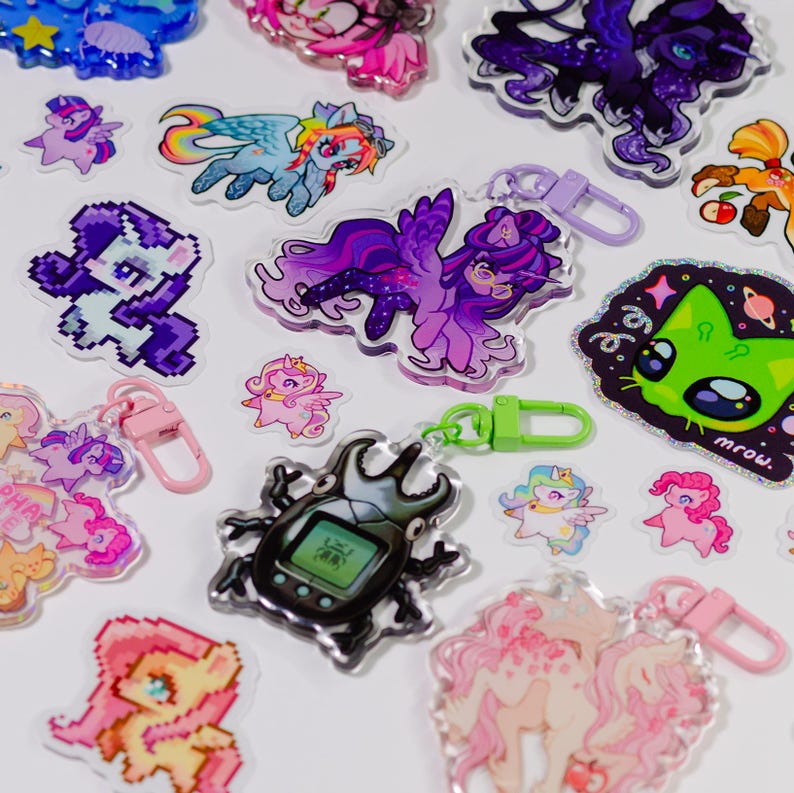 May include: A selection of vibrant acrylic keyrings and stickers showcasing cartoon characters and designs. The keyrings have metal clasps in various colours. The stickers include a green cat with the word "mrow" and a beetle with a digital screen.
