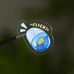 May include: A clear sticker featuring a white computer mouse with a blue circular area containing a small yellow and white clownfish. The text *CLICK!* is printed in yellow above the mouse, accompanied by two light blue motion lines.
