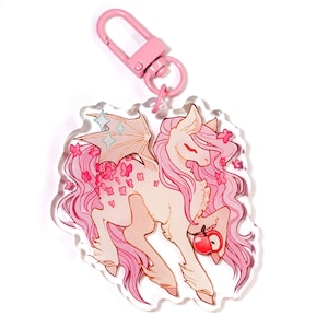 May include: A pink and white acrylic keychain featuring a cartoon pony with pink hair, wings, and floral accents. The pony is holding a red apple. The keychain has a pink clasp.