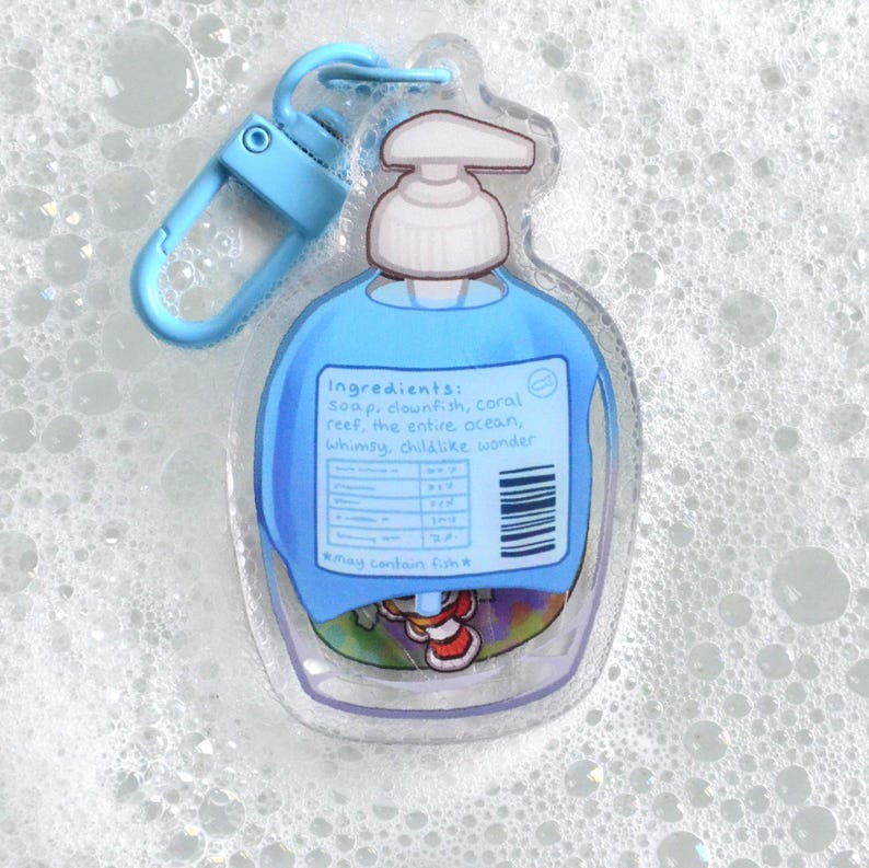 May include: A clear, soap-shaped keychain with a blue liquid design and a blue clip. The label reads "Ingredients: soap, clownfish, coral reef, the entire ocean, whimsy, childlike wonder." The keychain has a whimsical, cartoonish style.