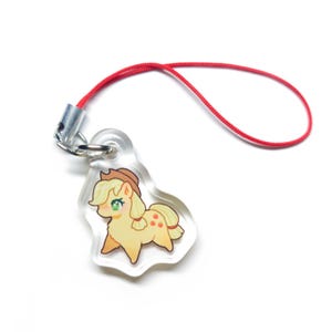 May include: A clear acrylic charm depicting a cartoon pony with a brown hat, yellow hair, and red apple markings. The charm is attached to a red cord and silver-coloured metal hardware. The charm is approximately 5 cm in length.