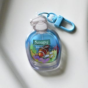 May include: A clear, plastic keychain shaped like a soap bottle with a blue liquid inside. The bottle has a white pump and a blue clip. The label reads "Soapy aquarium" with a cartoon clownfish and other fish.