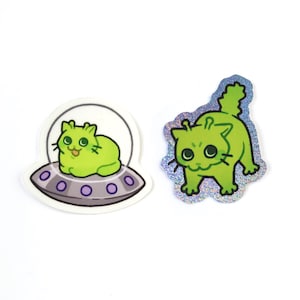 May include: Two stickers featuring green alien cats. One cat is inside a UFO, and the other is standing with a holographic border. The stickers have black whiskers and large eyes. The UFO has purple lights.