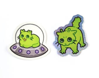 以下が含まれることがあります： 緑色の宇宙猫の2つのステッカー。1匹の猫はUFOの中にいて、もう1匹はホログラフィックの境界線で立っています。ステッカーには黒いひげと大きな目があります。UFOには紫色のライトがあります。