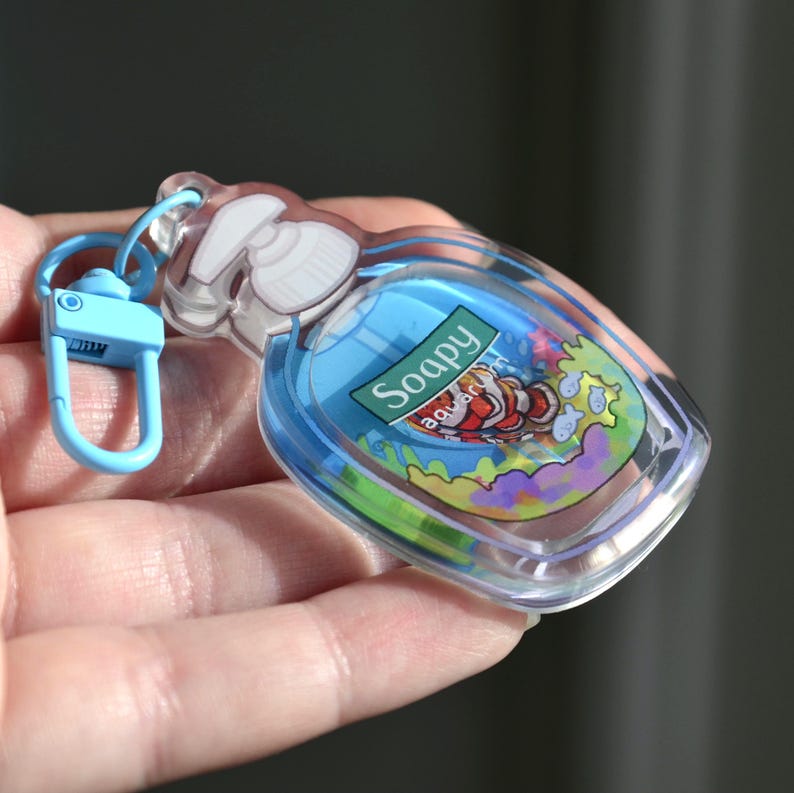 May include: A clear, soap-shaped keychain with a blue liquid interior and a cartoon aquarium scene. The keychain has a blue clasp and the text "Soapy Aquarium" on a green label. The design features a clownfish and colorful coral.