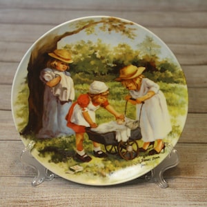 Puede incluir: Un plato de porcelana vintage con una escena pintada de tres niñas en un jardín. Las niñas llevan vestidos y sombreros. Una niña está empujando una muñeca en un cochecito. El plato está en un soporte blanco.