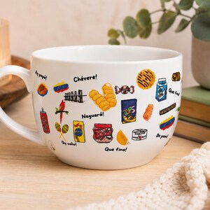 May include: White ceramic mug with a large handle, decorated with colorful illustrations of Venezuelan snacks and phrases. The mug features images of food items, a red bird, and the words "Ay pap&aacute;!" and "Ch&eacute;vere!"