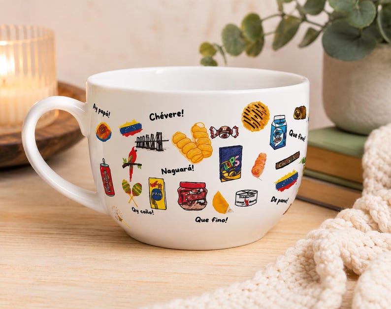 May include: White ceramic mug with a large handle, decorated with colorful illustrations of Venezuelan snacks and phrases. The mug features images of food items, a red bird, and the words "Ay pap&aacute;!" and "Ch&eacute;vere!"
