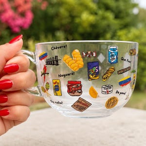 May include: A clear glass mug with a handle, decorated with colorful illustrations of Venezuelan food items and phrases. The mug is held by a hand with red nail polish, with a blurred green and pink background.
