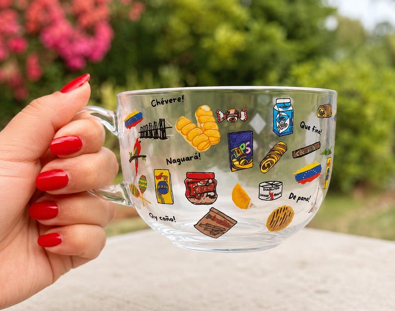 May include: A clear glass mug with a handle, decorated with colorful illustrations of Venezuelan food items and phrases. The mug is held by a hand with red nail polish, with a blurred green and pink background.