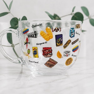 May include: Clear glass mug decorated with colorful Venezuelan food and cultural icons. The mug features images of snacks, flags, and phrases like "Ch&eacute;vere!" and "Naguar&aacute;!" A perfect gift for anyone who loves Venezuela.