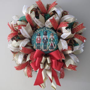 May include: A Christmas wreath decorated with red, white and gold ribbon. The wreath features a round, green and blue ornament with a picture of three nutcracker figurines and the text "Merry Christmas".