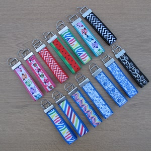 May include: A collection of 12 colourful keychains with various patterns, including owls, ladybirds, zigzags, and watermelon. The keychains are made of fabric and have silver metal rings.