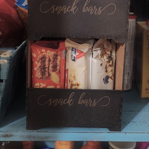 May include: Two black wooden snack bar organizers with the words "snack bars" written on the front. The organizers are filled with various snack bars, including Nature's Bakery Fig Bar, Atkins Peanut Butter Bars, and Clif Bars.