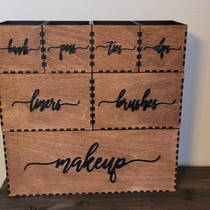 May include: A wooden makeup organizer with six compartments. The compartments are labeled "brushes", "liners", "lips", "eyes", "pins", and "bands". The bottom compartment is labeled "makeup".