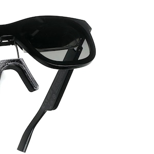 May include: Black sunglasses with a flexible, adjustable arm. The sunglasses have a dark gray lens and a black frame.