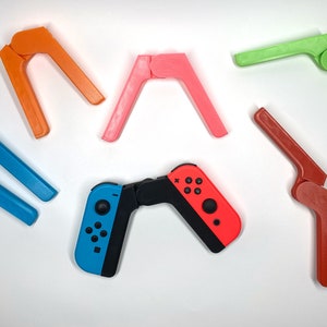May include: Five colorful plastic stands for a Nintendo Switch controller, in orange, pink, green, red, and blue. The stands are designed to prop up the controller for easier gaming.