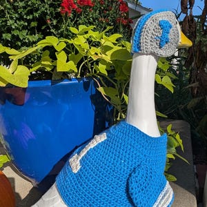 May include: A ceramic goose figurine wearing a handmade blue and gray crochet football jersey and hat. The jersey has a white stripe and the number 1. The hat also features the number 1.